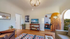 A spacious living room with a wooden floor, a large TV, a chandelier, and a view through an arched doorway.