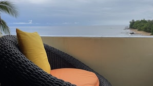 Ein Korbstuhl mit gelben und orangen Kissen, mit Blick auf einen Strand mit Palmen und einem bewölkten Himmel.