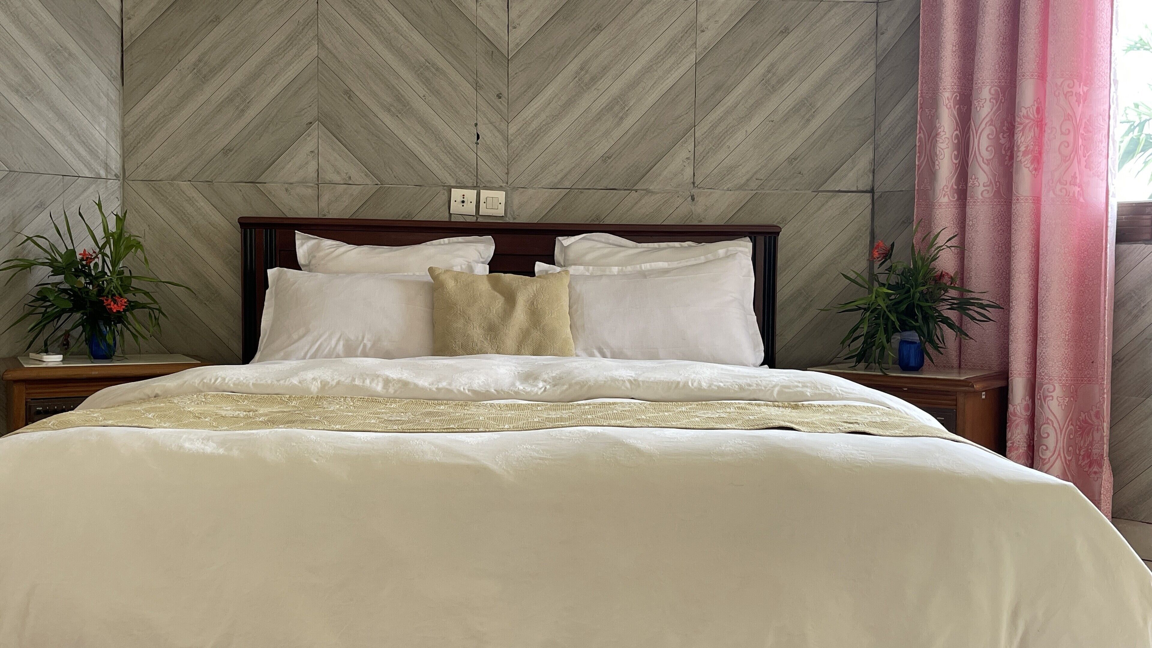 Una cama impecable con sábanas blancas y una manta color beige, flanqueada por dos mesitas de noche con plantas en macetas.