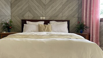 Una cama impecable con sábanas blancas y una manta color beige, flanqueada por dos mesitas de noche con plantas en macetas.