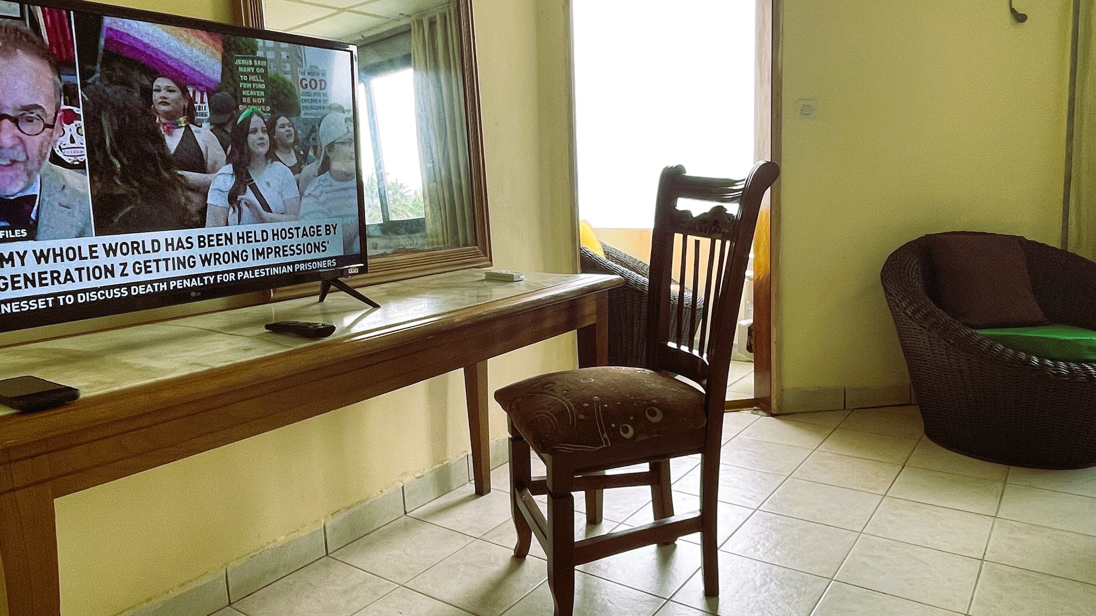 Une pièce équipée d’un téléviseur diffusant les actualités, d’un bureau en bois, d’une chaise et d’un canapé en osier.