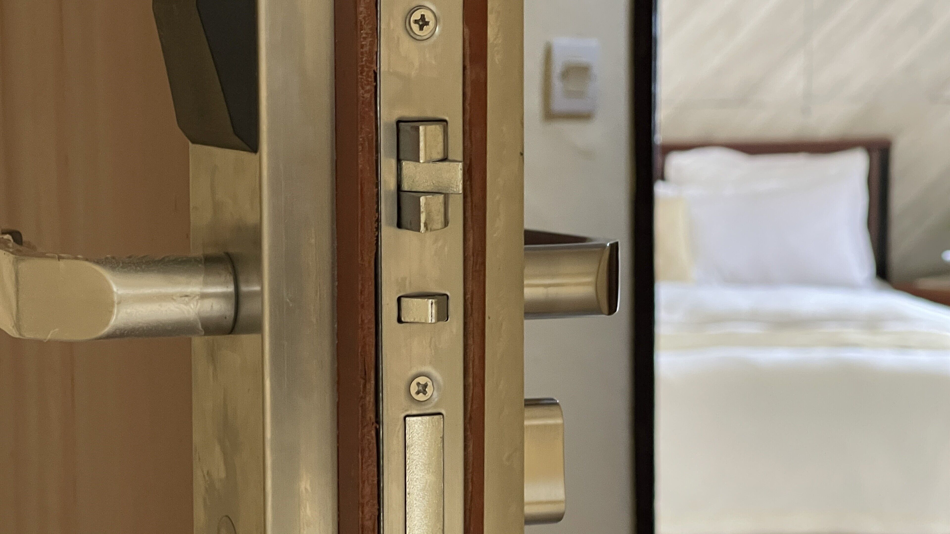 A hotel room door with a metallic handle and lock, a visible bed with white bedding, and a small wall-mounted light fixture.