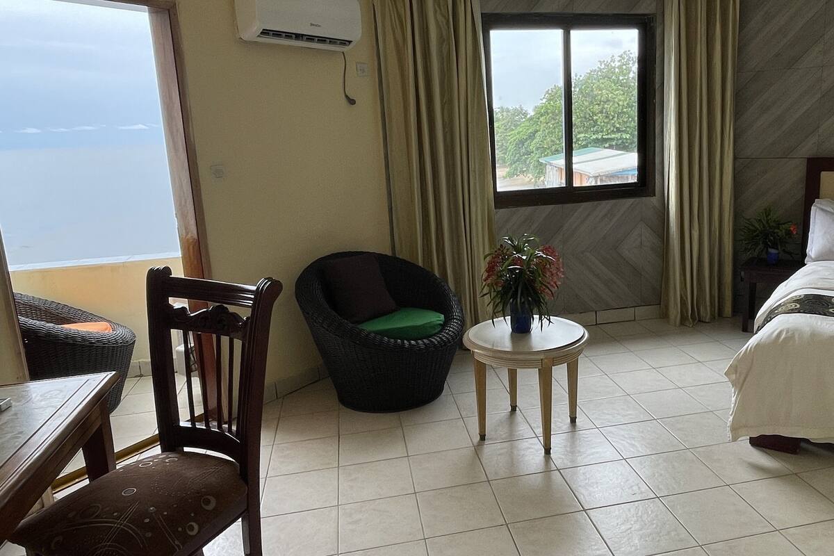 Une chambre d’hôtel avec un lit, une chaise, une petite table sur laquelle se trouve un vase, et une fenêtre avec des rideaux.