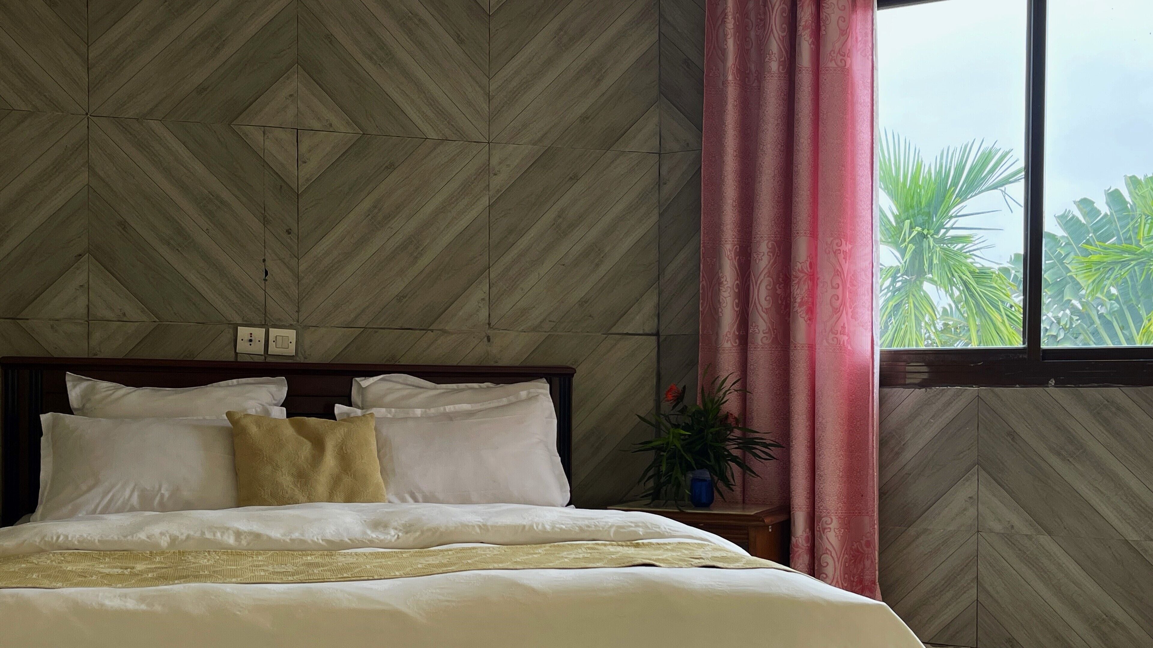 A hotel room with a herringbone-patterned wall, a wooden headboard, a bed with white and beige bedding, a red curtain, and a window with a view of palm trees.