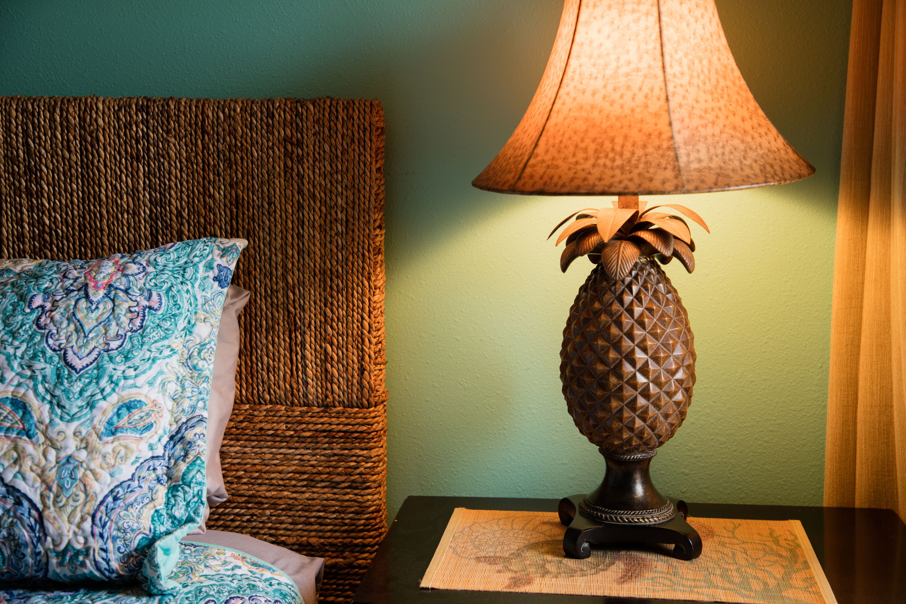 A bed with a patterned pillow and a woven headboard, a table with a pineapple-shaped lamp, and a green wall.