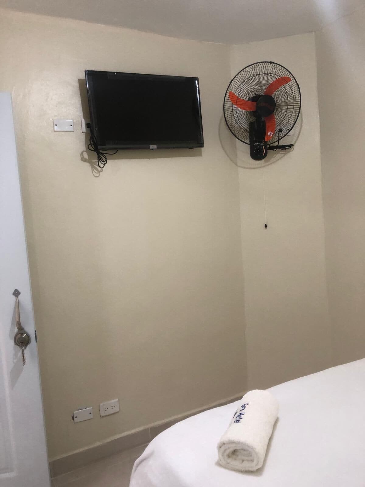 Una habitación con cama, televisor montado en la pared, ventilador de pie y una puerta con cerradura de llave.