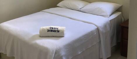 A single bed with white bedding and pillows, a small wooden nightstand, and a window with curtains.