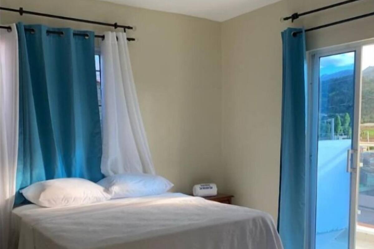 Een slaapkamer met een bed, gordijnen, een nachtkastje en uitzicht op een balkon.