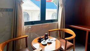 A room with a round wooden table, two chairs, a window with curtains, and a view of buildings with blue roofs. - Cao Lanh Hotel (My Tho)