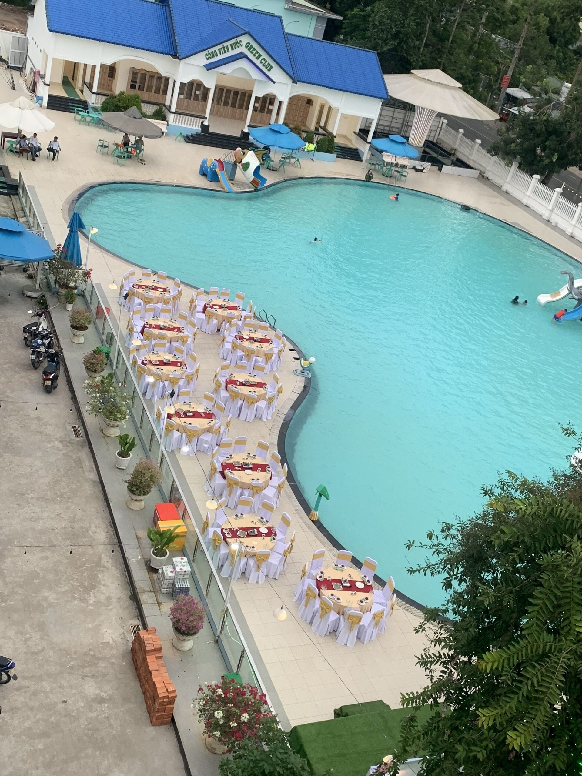 Zona de piscina con varias mesas y sillas redondas, edificio con tejado azul y área de natación cercada.