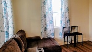 A living room with a brown sofa, a black chair, and a window with floral curtains. - Private Guest Suite (Visalia)