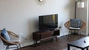 Ein Wohnzimmer mit Flachbildfernseher auf Holzfuß, zwei Korbstühle mit Kissen, einer Stehlampe und einer runden Wanddekoration.