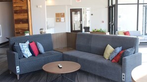 Eine moderne Lobby mit einem grauen Ecksofa, einem runden Holztisch und einer Glastür, die nach draußen führt.