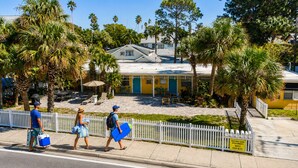 A beachfront property with a white picket fence, palm trees, and a building with a yellow facade. - Miami & Me Oasis: Dive into Beach Bliss, King Size Comfort, and Gulfside Glam! (Indian Rocks Beach)