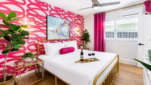 Hotelkamer met een bed, een plafondventilator, een raam met gordijnen en een muur met een abstract roze patroon.