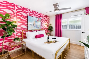 A hotel room with a bed, a ceiling fan, a window with curtains, and a wall with a pink abstract pattern. - Miami & Me Oasis: Dive into Beach Bliss, King Size Comfort, and Gulfside Glam! (Indian Rocks Beach)