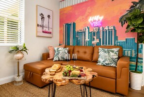 A modern living room with a brown leather sofa, a wooden coffee table with food, a cityscape painting, and a neon sign.