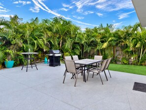 Un patio exterior con una parrilla, mesa y sillas, rodeado de abundante vegetación.