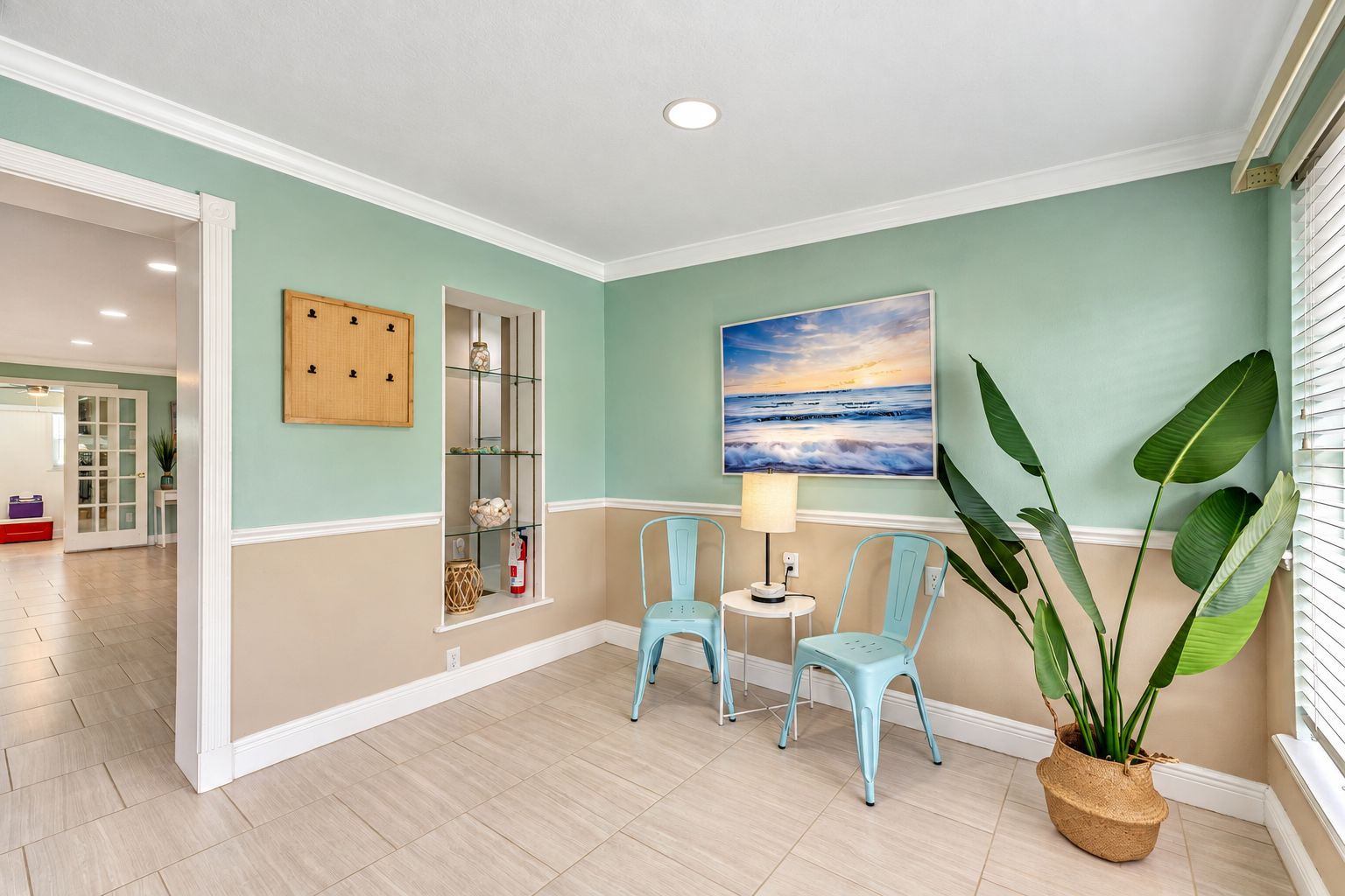 Un couloir avec un mur de couleur turquoise, un meuble en bois, une étagère en verre, deux chaises, une table avec une lampe et une grande plante.
