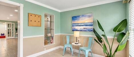 Un couloir avec un mur de couleur turquoise, un meuble en bois, une étagère en verre, deux chaises, une table avec une lampe et une grande plante.