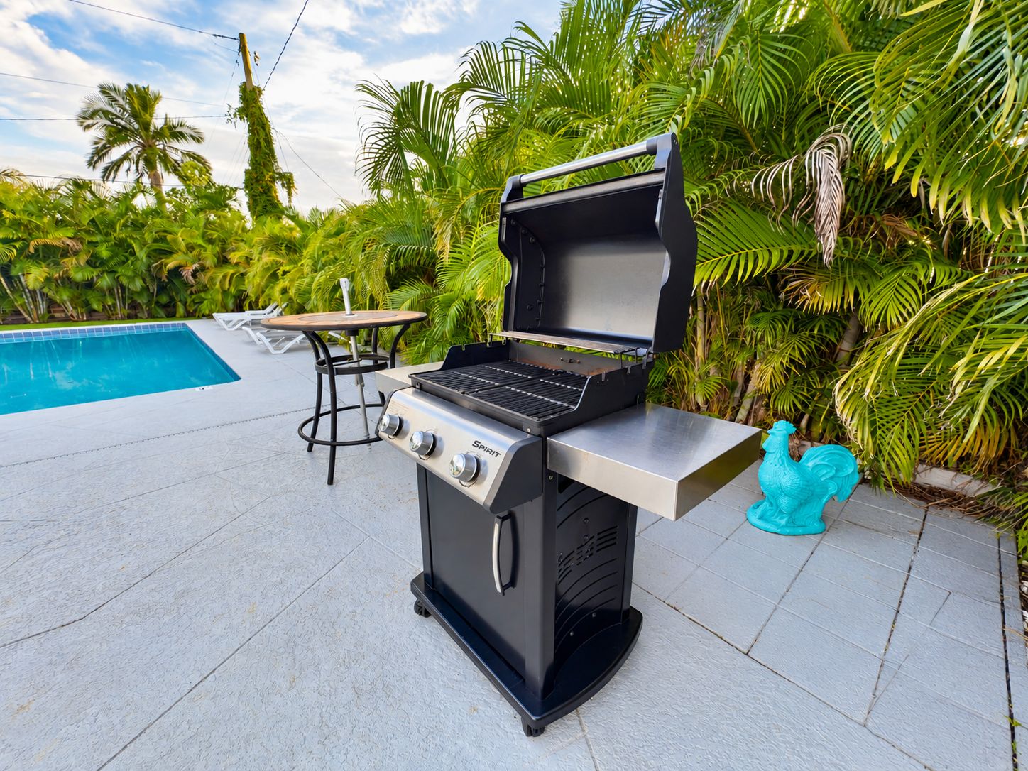 Churrasqueira ao ar livre com mesa, cadeiras, piscina e plantas tropicais.