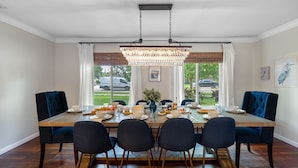 Comedor con mesa de madera dispuesta para seis personas, rodeada de sillas tapizadas oscuras. Un gran candelabro cuelga del techo, y la habitación tiene ventanas amplias con cortinas blancas.