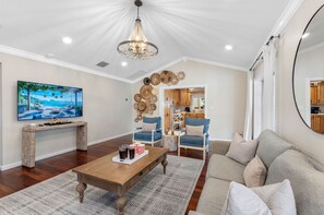 A spacious living room with a wooden coffee table, blue armchairs, a beige sofa, and a large flat-screen TV mounted on the wall.