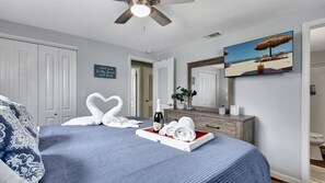 Une chambre à coucher comprenant un lit, une commode, un miroir, un ventilateur de plafond, un téléviseur fixé au mur et un tableau sur le thème de la plage.