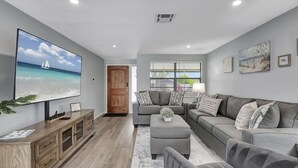 Een moderne woonkamer met een grote flatscreen televisie, grijze banken, een houten kast en uitzicht op de oceaan door een raam.