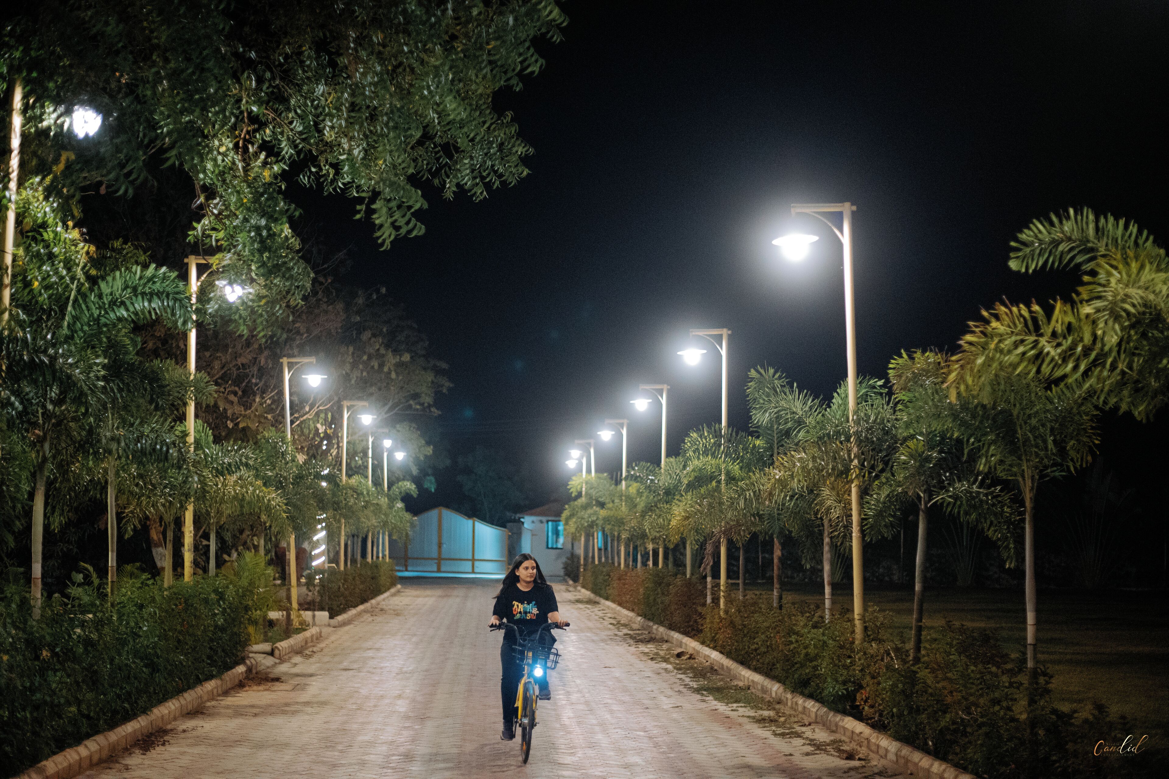 A person riding a bicycle on a paved path at night, flanked by streetlights and lined with trees.