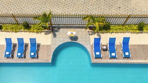 Vista aerea di una zona piscina con un elemento d'acqua centrale, circondata da poltrone da relax blu e palme.