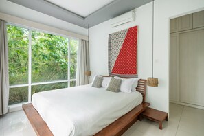 A bedroom with a large window, a bed with patterned pillows, a wall-mounted air conditioner, and a wooden bench.