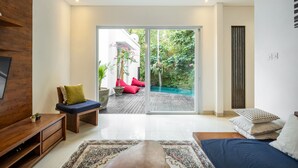 Un salon moderne avec un meuble TV en bois, un canapé avec des coussins, un tapis à motifs et une grande fenêtre donnant sur une piscine et la verdure environnante.