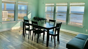 Uma área de refeições com mesa e cadeiras, grandes janelas com vista para a praia e oceano, e piso de madeira.