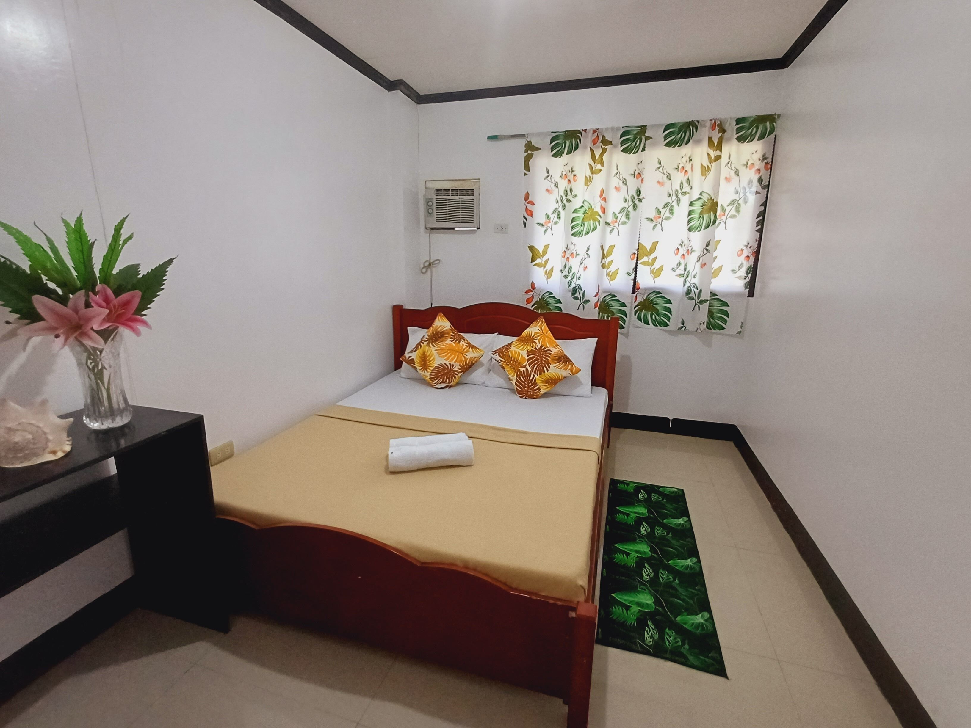 Een slaapkamer met een bed, een nachtkastje, een vaas met bloemen en een raam met gordijnen.