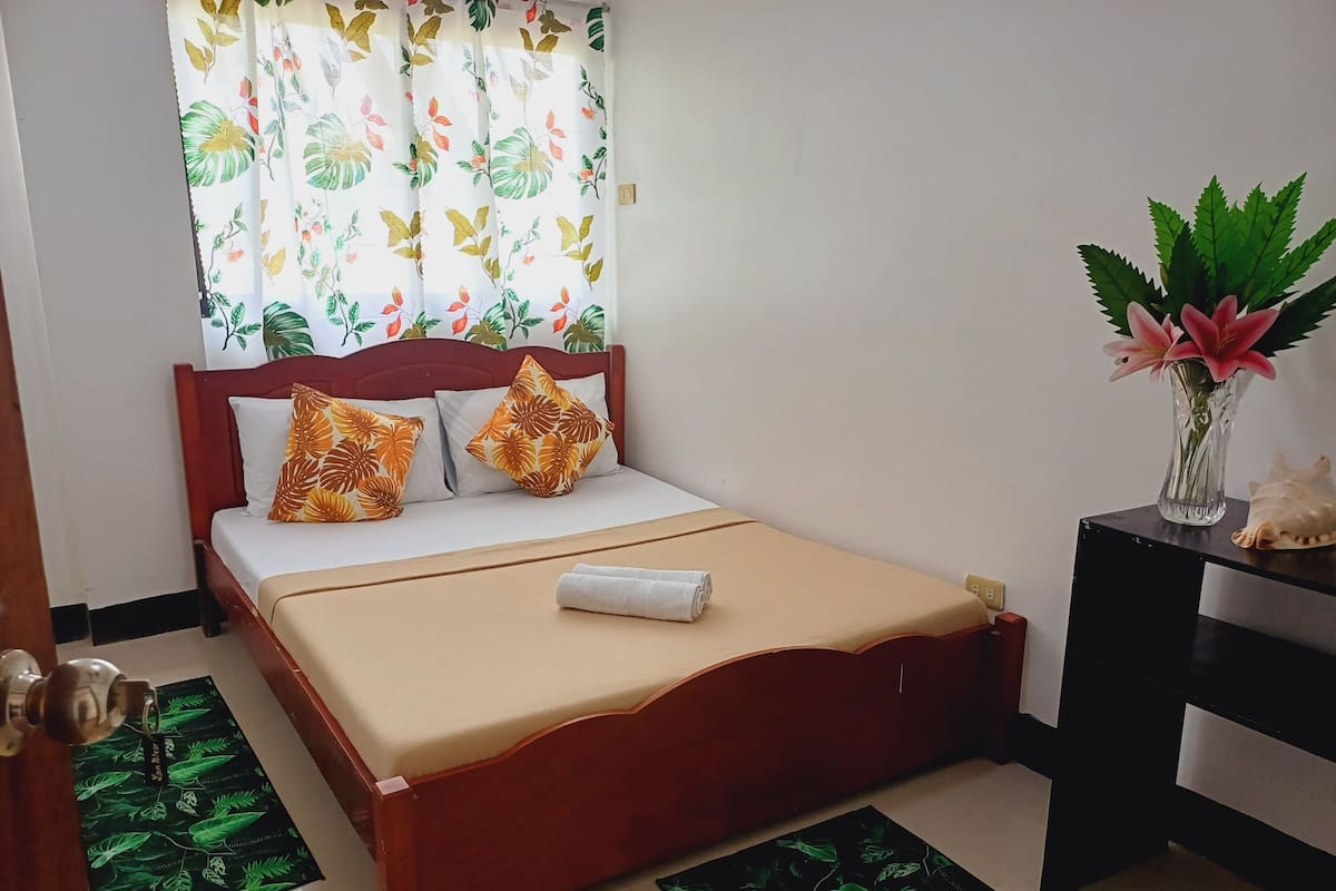 A neatly made bed with a beige bedspread and two decorative pillows, a wooden headboard, and a nightstand with a vase of flowers.