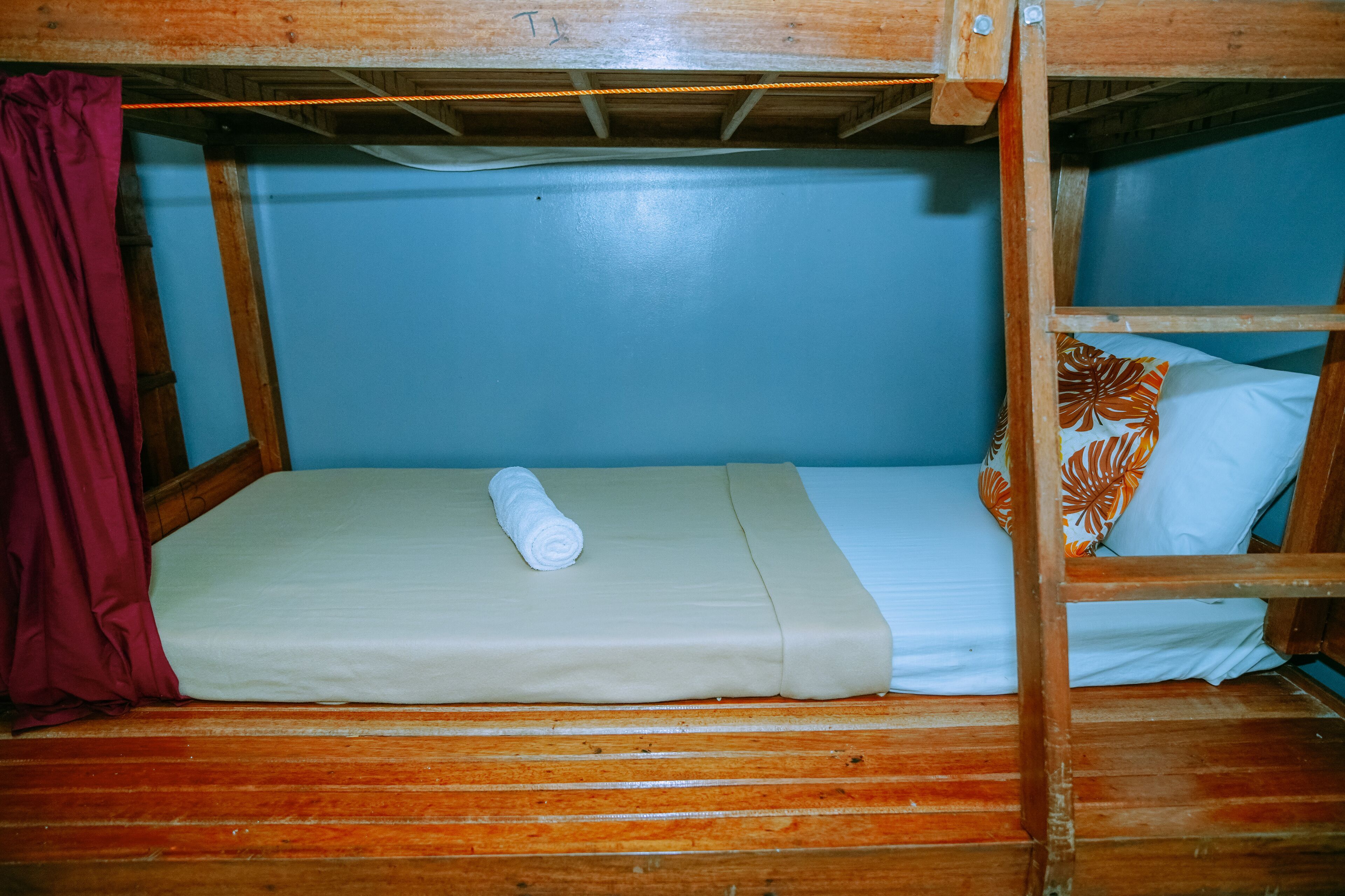 A bunk bed with a rolled towel on the lower bunk, a pillow with a leaf pattern, and a maroon curtain.