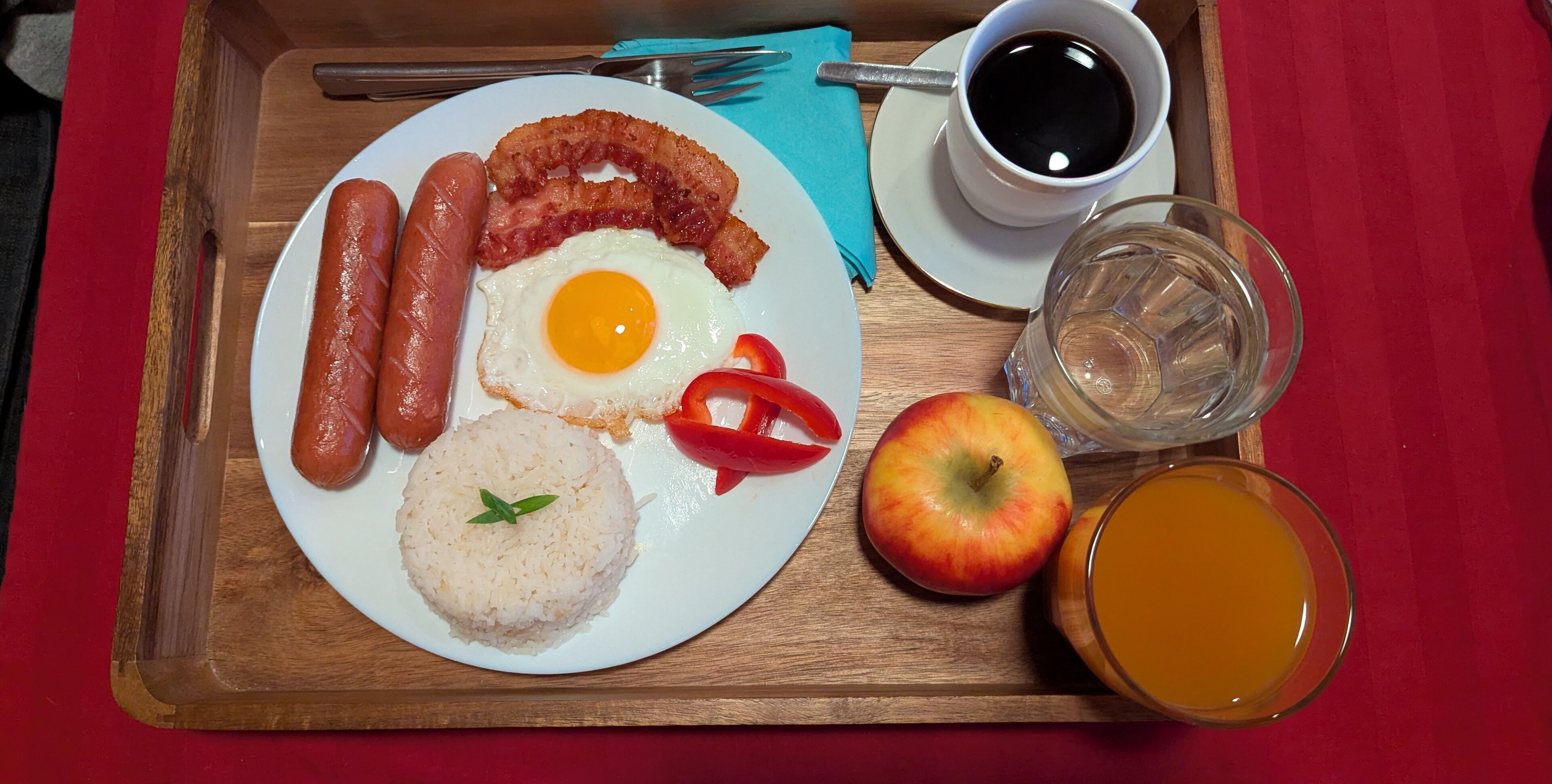 A breakfast tray with sausages, bacon, an egg, rice, an apple, and a glass of orange juice.