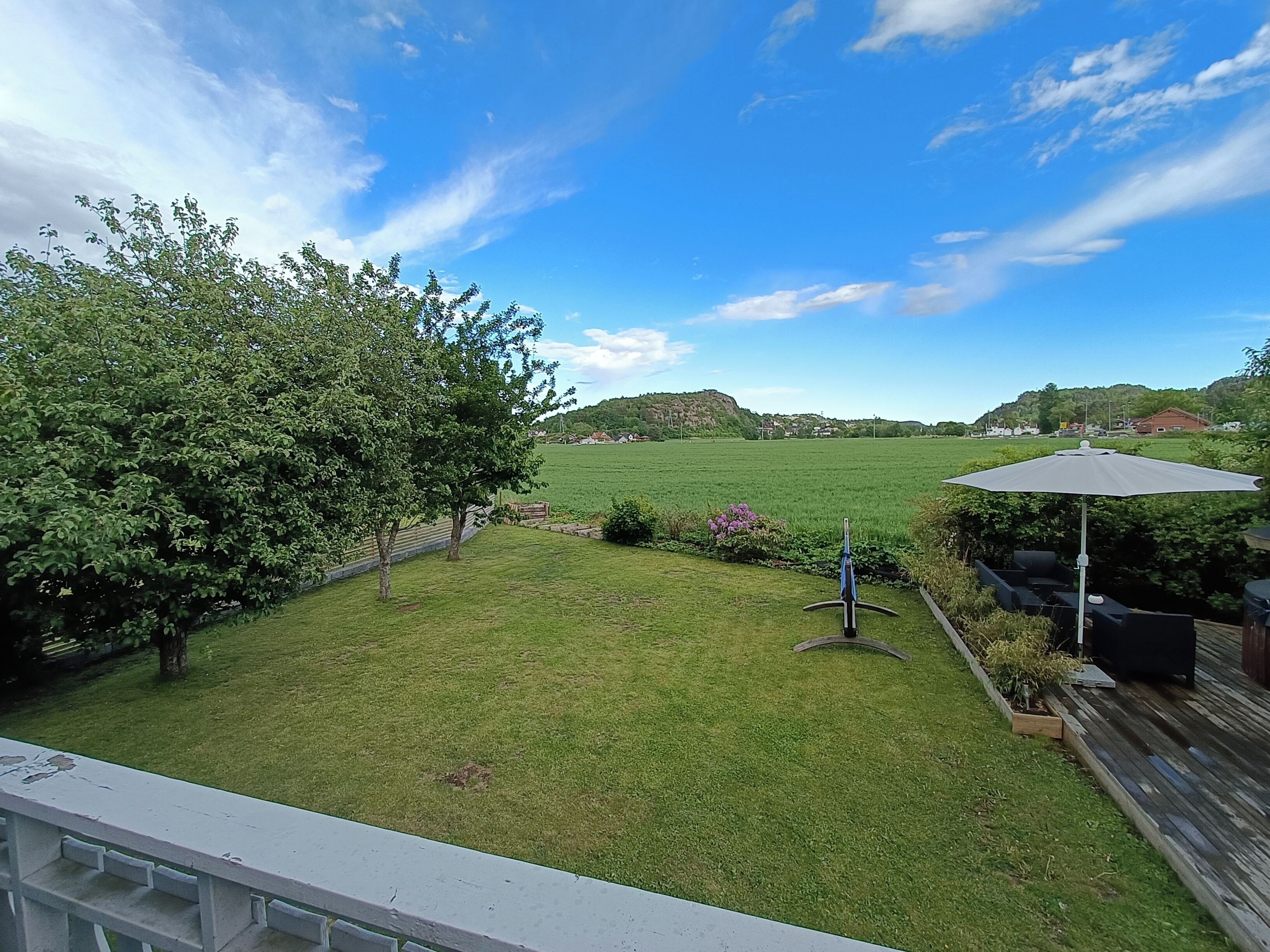 A backyard with a grassy lawn, a tree, a patio with outdoor furniture, and a view of a distant hill.