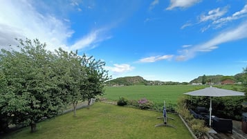 A backyard with a grassy lawn, a tree, a patio with outdoor furniture, and a view of a distant hill.
