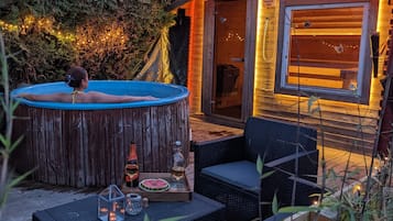 A hot tub in a wooden cabin with a glass door, surrounded by greenery and string lights.