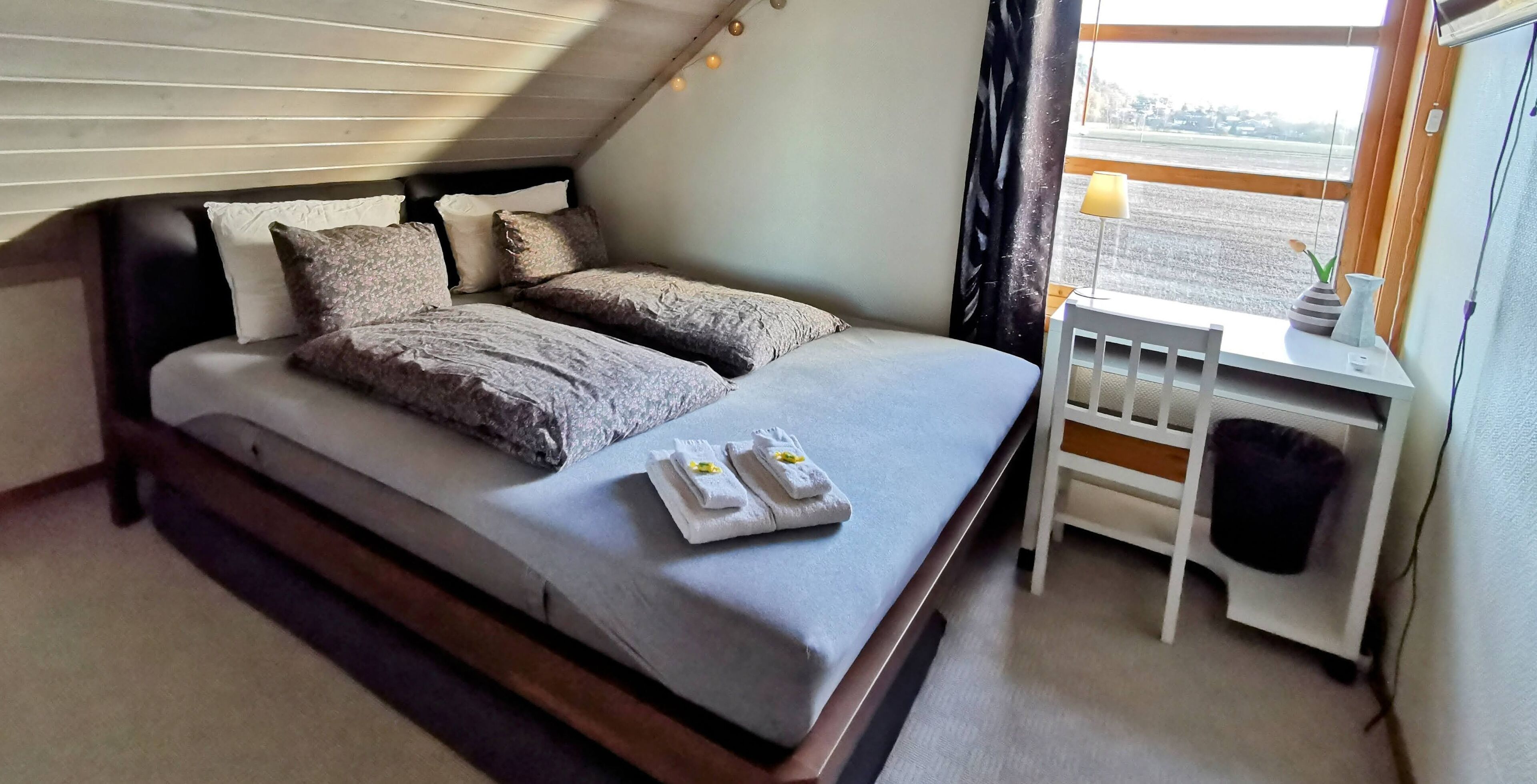 A neatly made bed with pillows and towels, a small bedside table with a lamp, and a view of the outside through a window.