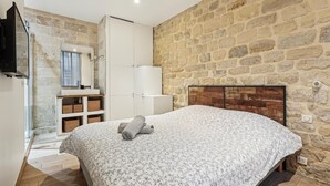A bedroom with a stone wall, a wooden headboard, a bed with a floral bedspread, a white wardrobe, and a bathroom with a stone wall and a mirror.
