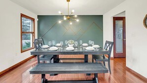 Comedor con una mesa de madera oscura dispuesta para seis comensales, una lámpara moderna y una pared de acento verde.