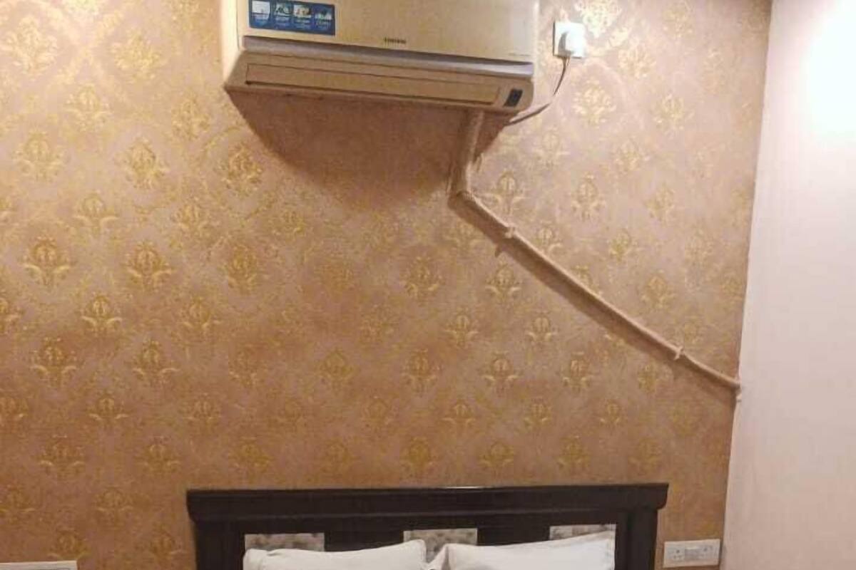A bedroom with a bed, white pillows, a nightstand, a wall-mounted air conditioner, and patterned wallpaper.