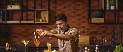 Un bar con un cantinero preparando bebidas, una selección de quesos y varias botellas en la barra.