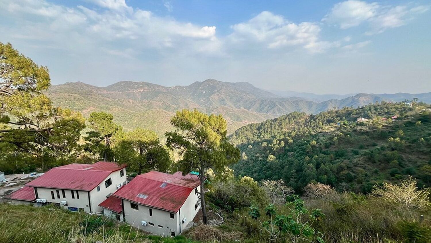 A rural landscape with a white building with a red roof, surrounded by trees and hills in the background.