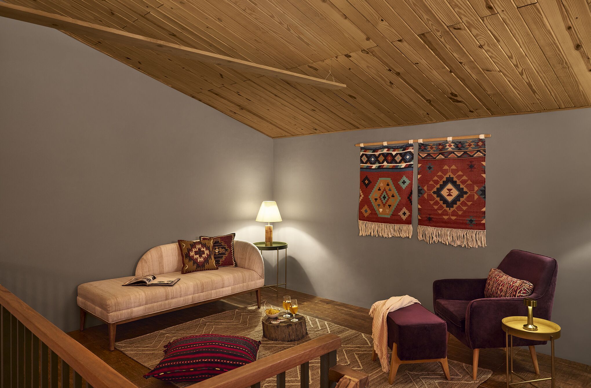 A cozy living room with a wooden ceiling, a sofa, a chair, a lamp, and two wall hangings.