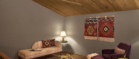 A cozy living room with a wooden ceiling, a sofa, a chair, a lamp, and two wall hangings.