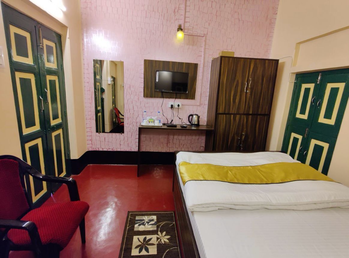 Une chambre d’hôtel avec un lit, un fauteuil rouge, une armoire en bois, un téléviseur fixé au mur et une petite table avec une bouilloire et des bouteilles.